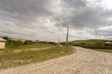 Bir kaç evi ve ortasında bir direği olan kırsal bir yol. Gökyüzü bulutlu ve yol kirli.