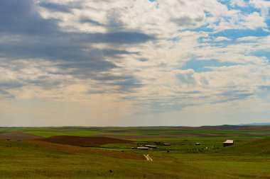 Geniş, açık bir alan, birkaç bina uzakta. Gökyüzü bulutlu ve güneş bulutların arasında parlıyor.