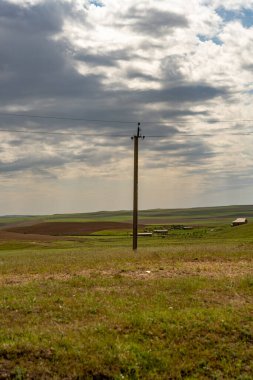 Tepesinde bulutlu bir gökyüzü olan bir tarlada uzun bir elektrik direği duruyor. Ortalık sakin ve sakin. Etrafta ne insan ne de hayvan var.