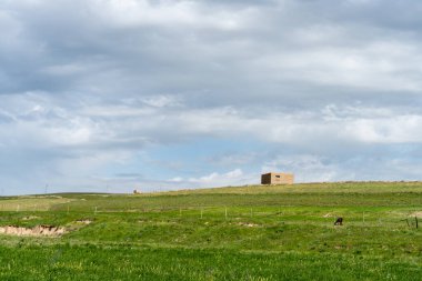 Tarladaki bir tepede büyük, boş bir bina duruyor. Gökyüzü bulutlu ve çimenler yeşil