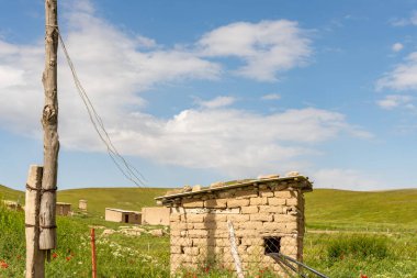 Önünde çit olan küçük bir kulübe. Gökyüzü mavi ve bazı bulutlar var.