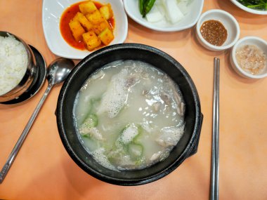 Bir kase çorba bir kaşık ve bir kase pirinçle masada oturur. Çorba beyaz ve içinde yeşil bir sebze var.