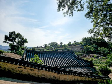 Önünde eğimli bir çatı ve bir ağaç olan bir ev. Ev ağaçlar ve tepelerle çevrili.