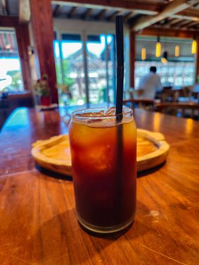 İçinde pipet olan bir bardak buzlu kahve ahşap bir masanın üzerinde duruyor. Masa sandalyelerle çevrilidir ve arka planda bir kişi görünür. Sahne rahat ve rahat bir atmosfer sunuyor.