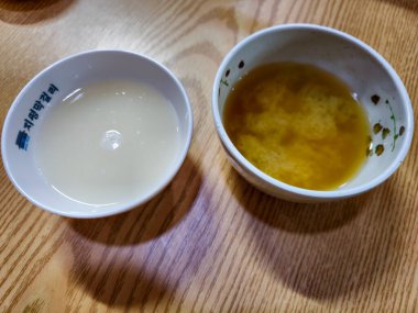 İki kase yemek, biri beyaz, biri sarı, tahta bir masaya otur. Kaseler farklı yemek türleriyle doludur ve masa ahşaptan yapılmıştır. Sıcaklık ve rahatlık kavramı