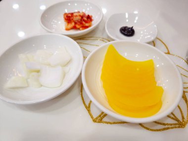 Üzerinde dört kase yiyecek olan beyaz bir masa. Kâselerde havuç, soğan ve turşu da dahil olmak üzere farklı yemek türleri yer alıyor. Masa yemek için hazır ve yemek taze ve iştah açıcı görünüyor.