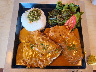 Pirinç, et ve sebzeli bir tabak yemek. Tabak kare ve siyah. Yemek lezzetli ve iştah açıcı görünüyor.