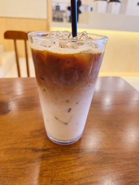 Bir bardak buzlu kahve ahşap bir masanın üzerinde duruyor. Kahve espresso ve sütün karışımı, ve buz eriyor, köpüklü bir doku oluşturuyor. Sahne rahat ve rahat.