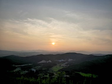 Bir dağ sırasının üzerinde güzel bir gün batımı. Gökyüzü bulutlarla dolu ve güneş uzaklardan batıyor. Dağlar ağaçlarla kaplı ve manzara sakin ve huzurlu.