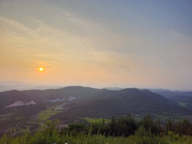Güneş dağların üzerinde batıyor, manzaranın üzerine sıcak bir parıltı saçıyor. Gökyüzü bulutlarla dolu. Huzurlu ve huzurlu bir atmosfer yaratıyor.