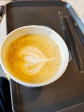 Üstünde kalp şekli olan bir fincan kahve istiyorum. Kahve bir kağıt bardakta ve bir tepside oturuyor.