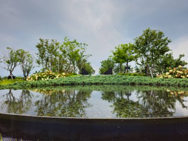 Ağaçların yansıması ve birkaç sandalyenin olduğu bir gölet. Gökyüzü bulutlu ve su sakin.