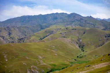 Üzerinde açık mavi gökyüzü olan güzel, yemyeşil bir yamaç. Arka plandaki dağlar manzaranın manzaralı güzelliğine eklenir