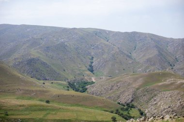İçinden bir nehir akan güzel bir dağ sırası. Manzara yemyeşil ve yemyeşil, tepeleri ağaçlar ve otlar kaplıyor. Gökyüzü açık ve mavidir ve güneş ışıl ışıl parlar.