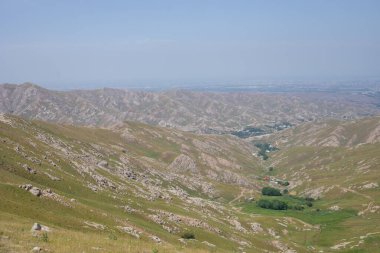 Aralarında vadi olan bir dağ sırası. Vadi yeşildir ve birkaç ağacı vardır. Gökyüzü açık ve güneş parlıyor.