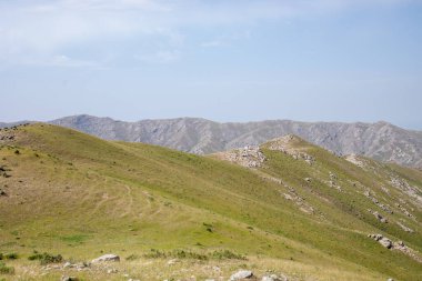 Arka planda açık mavi gökyüzü olan bir dağ sırası. Dağlar çimen ve kayalarla kaplıdır.