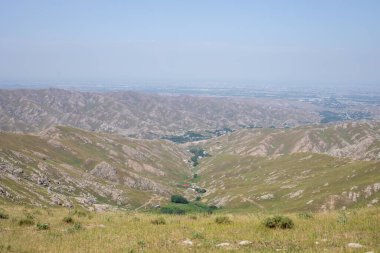 Aralarında vadi olan bir dağ sırası. Vadi yeşildir ve birkaç ağacı vardır. Gökyüzü açık ve güneş parlıyor.