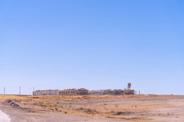 Issız bir çöl manzarası ve birkaç bina uzakta. Gökyüzü açık ve mavi. Sahne yalnızlık ve boşluktur.