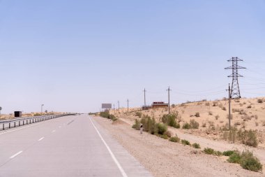 Arka planda birkaç ağaç ve reklam panosu olan bir yol. Gökyüzü açık ve güneş parlıyor.