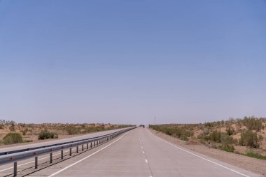 Üzerinde mavi bir gökyüzü olan uzun, boş bir yol. Yol metal bir çitle kaplı.