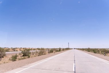 Üzerinde açık mavi gökyüzü olan uzun, boş bir yol. Yol ağaçlarla kaplı ve yolun ortasında bir direk var.