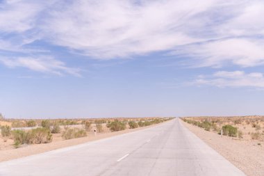 Üzerinde açık mavi gökyüzü olan uzun, boş bir yol. Yol bir çöl manzarasıyla çevrilidir.