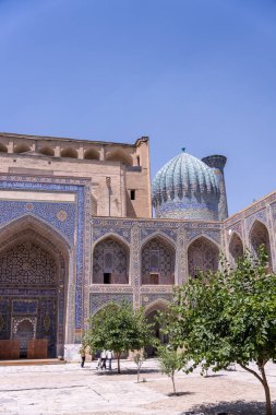 Büyük bir bina ve tepesinde kubbe olan bir avlu. Avlu ağaçlar ve insanlarla dolu.