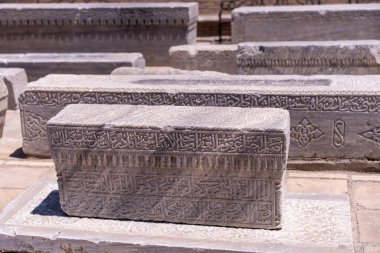 Üzerinde Arapça yazı olan bir taş levha yerde duruyor. Levha diğer taş levhalarla çevrili, bazılarında da yazı var. Bu sahne bir tarih hissi veriyor.