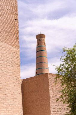 Üzerinde mavi çizgili uzun bir kule bir tuğla binanın önünde. Gökyüzü mavi ve güneş parlıyor.
