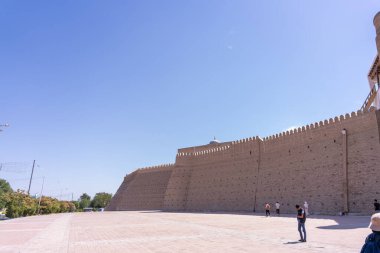 Bir grup insan büyük bir taş duvarın önünde duruyor. Gökyüzü açık ve mavi, ve arka planda park etmiş birkaç araba var.