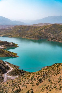 Kenarları boyunca uzanan bir yolu olan güzel bir dağ gölü. Su koyu mavi renktedir ve arkaplandaki dağlar yeşil ve kahverengi karışımıdır. Huzurlu ve huzurlu bir sahne.