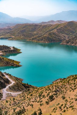 Arkasında dağlar olan güzel bir göl. Su mavi ve sakindir. Dağlar ağaçlarla örtülmüş, gökyüzü ise berrak.