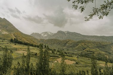 Arka planda bulutlu bir gökyüzü olan bir dağ sırası. Dağlar ağaçlarla çevrili, gök de bulutlarla çevrili.