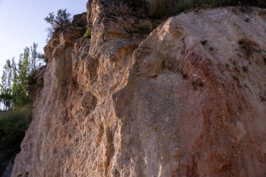 Arka planda birkaç ağaç olan kayalık bir uçurum. Uçurum kahverengidir ve bir sürü kayası vardır.