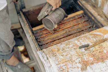 Bir insan ahşap tepsiden bir şey çıkarmak için elek kullanıyor. Tepsi kir içinde ve kişi gri pantolon giyiyor.