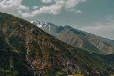 Dağlar yeşil ağaçlar ve kahverengi kayalarla kaplıdır. Gökyüzü bulutlu ve güneş parlıyor