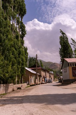 Toprak yolu ve birkaç evi olan küçük bir kasaba. Evler ahşaptan yapılmış ve kırsal bir havası var. Yol ağaçlarla kaplı ve yan tarafa park edilmiş birkaç araba var. Gökyüzü bulutlu.