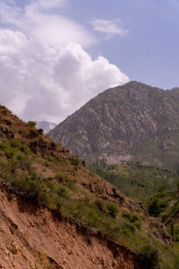 Arka planda bulutlu bir gökyüzü olan bir dağ sırası. Dağlar yeşil bitki örtüsüyle kaplıdır ve gökyüzü mavi ve beyaz bulutların karışımıdır.