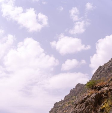 Gökyüzü bulutlu ve güneş bulutların arasında parlıyor. Bulutlar beyaz ve kabarık, ve gökyüzü açık mavi