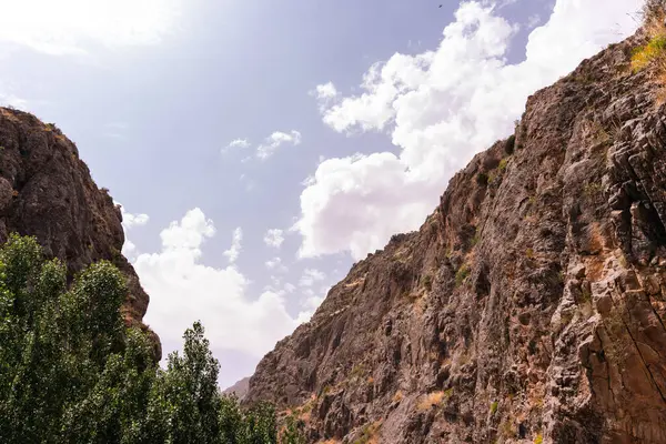 Gökyüzü açık ve mavidir. Birkaç bulut etrafa saçılmıştır. Arkadaki dağlar ağaçlar ve kayalarla kaplıdır.