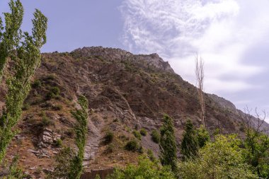 Arkasında bulutlu bir gökyüzü olan bir dağ. Dağ ağaçlar ve kayalarla kaplı. Gökyüzü mavi ve beyaz bulutların karışımıdır.