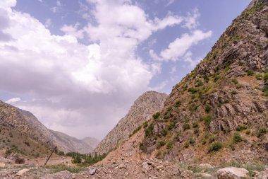 Arka planda bulutlu bir gökyüzü olan bir dağ sırası. Gökyüzü çoğunlukla mavi ve bazı bulutlar etrafa saçılmış. Dağlar ağaçlar ve kayalarla kaplı, manzarayı engebeli kılıyor.