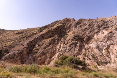 Üzerinde birkaç ağaç olan kayalık bir dağ. Gökyüzü açık ve mavi. Dağ çorak ve kayalık, bitki örtüsü yok.
