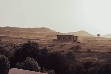 Kuru ve çorak bir arazide küçük bir ev bir yamaçtadır. Ev ağaçlarla çevrili ve arka planda bir çit var. Sahne sessiz ve huzurlu.