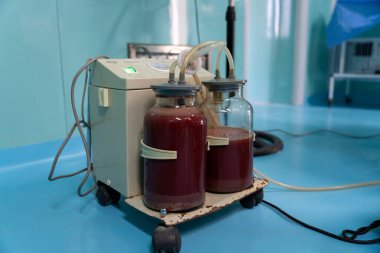 Üzerine iki tüp bağlanmış tıbbi bir cihaz. Tüpler kırmızı sıvıyla dolu. Cihaz tekerleklerin üzerinde ve mavi bir yüzeye yerleştirilmiş.