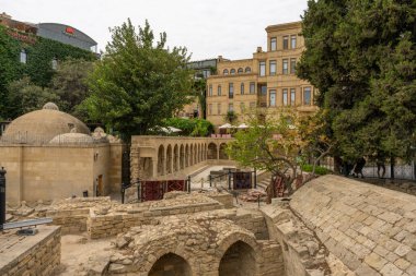 Arka planda bina olan bir avlu. Avlu taştan bir duvarla çevrili ve ortasında bir çeşme var.