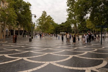 Kalabalık bir cadde, etrafta insanlar dolaşıyor ve arka planda büyük bir ağaç var. Manzara canlı ve hareketli.