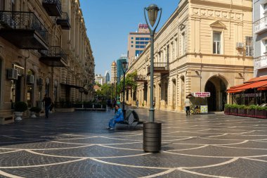 Mavi gömlekli bir adam şehir meydanında bir bankta oturuyor. Meydan boş ve binalar eski.