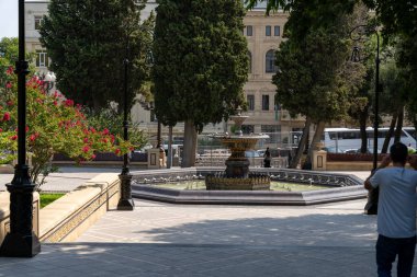 Bir adam parkta bir fıskiyenin fotoğrafını çekiyor. Çeşme ağaçlar ve banklarla çevrili ve yakınlarda park edilmiş birkaç araba var. Huzurlu ve huzurlu bir sahne.