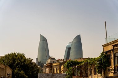 İki uzun bina ve büyük bir ağacı olan bir şehir manzarası. Binalar modern ve benzersiz bir tasarıma sahipken, ağaç kentsel çevrede bir doğa hissi sağlıyor. Sahne kontrastlardan biridir